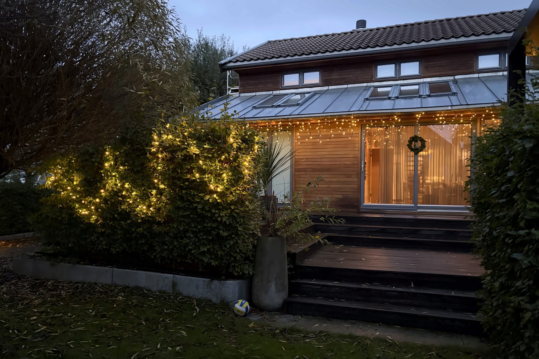 Bakside av et tre- og glasshus i blåtimen, med istapplys langs takkanten, krans på døren og en lysende hekk til venstre