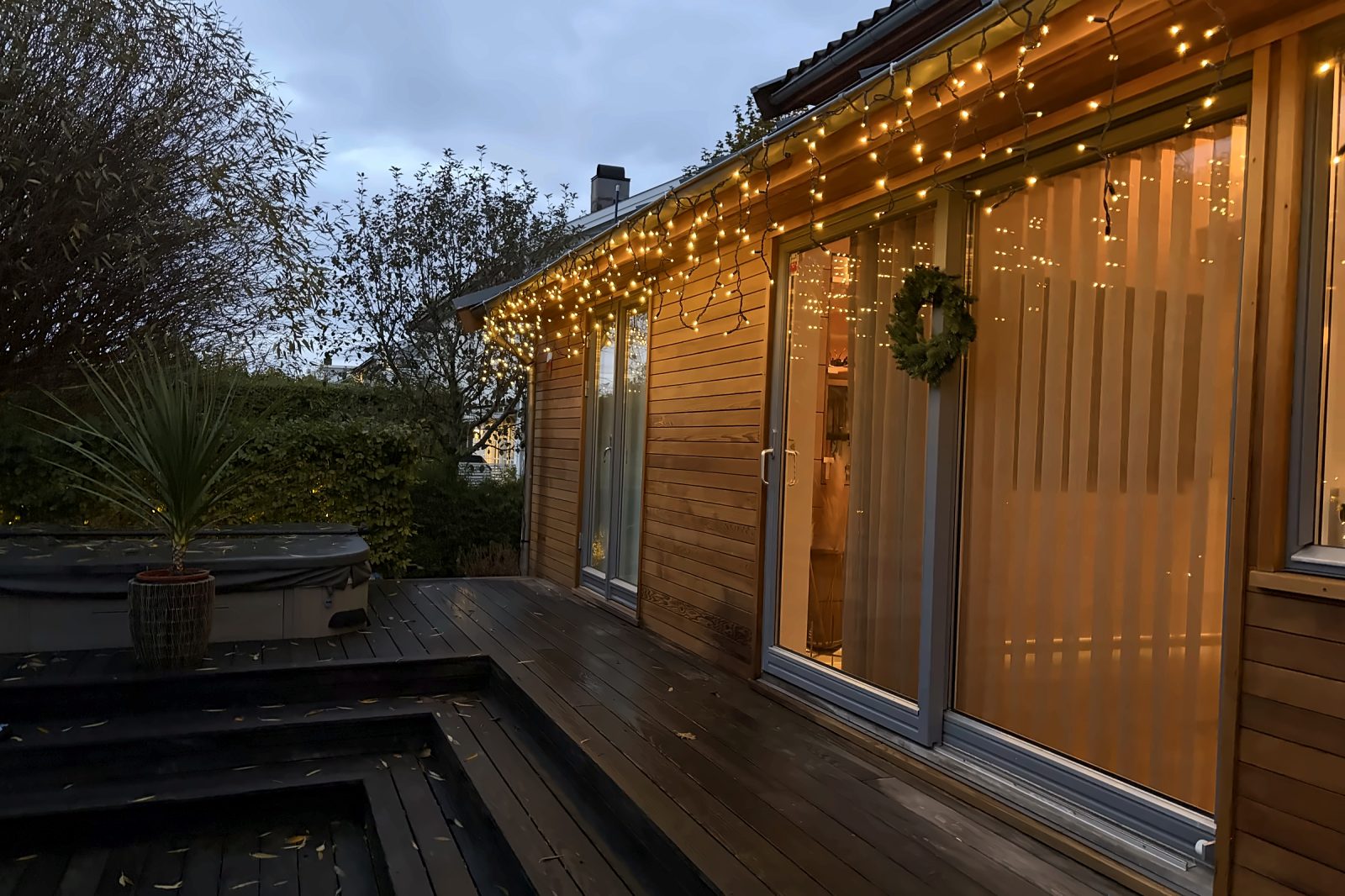 Terrasse med istapplys langs taket, krans på glassdøren og fuktig tredekke i blåtimen